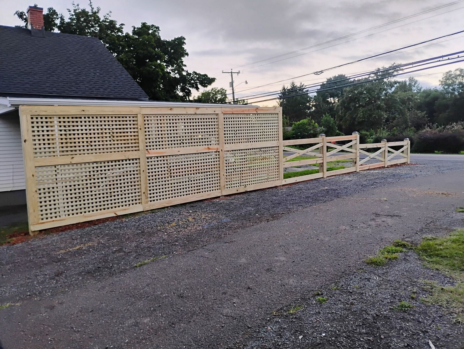 Wooden fence with lattice and cross-buck sections along a gravel driveway, next to a building.