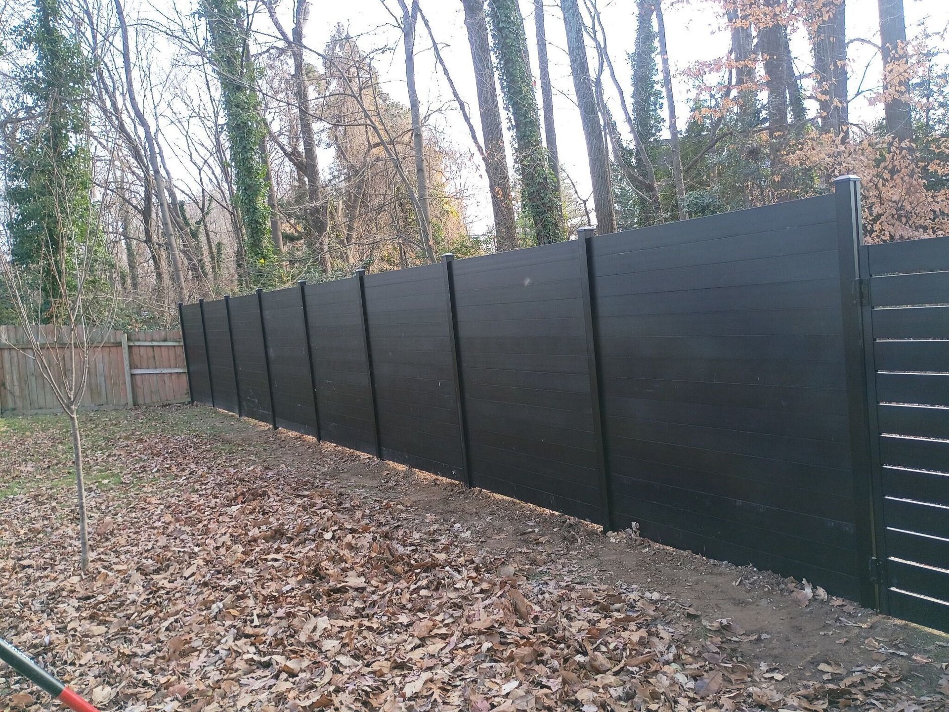 Black privacy fence in a backyard with fallen leaves and trees in the background.