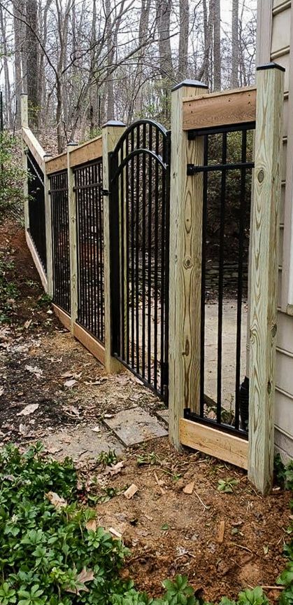 Black iron gate and fence with wooden frame in a wooded area.