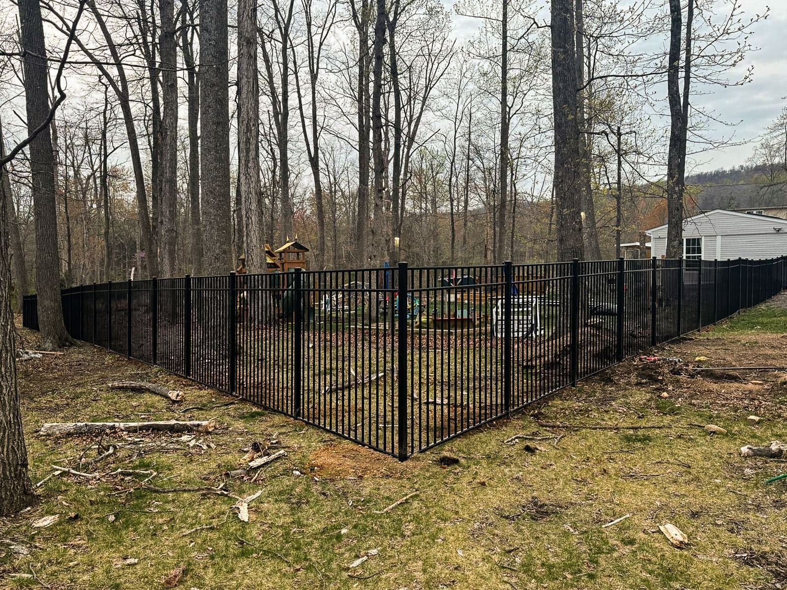Black metal fence surrounds a grassy yard in a wooded area with bare trees.