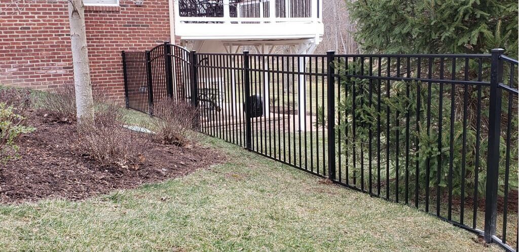 Black metal fence surrounding a yard with grass and bushes, a tree, and a brick building.