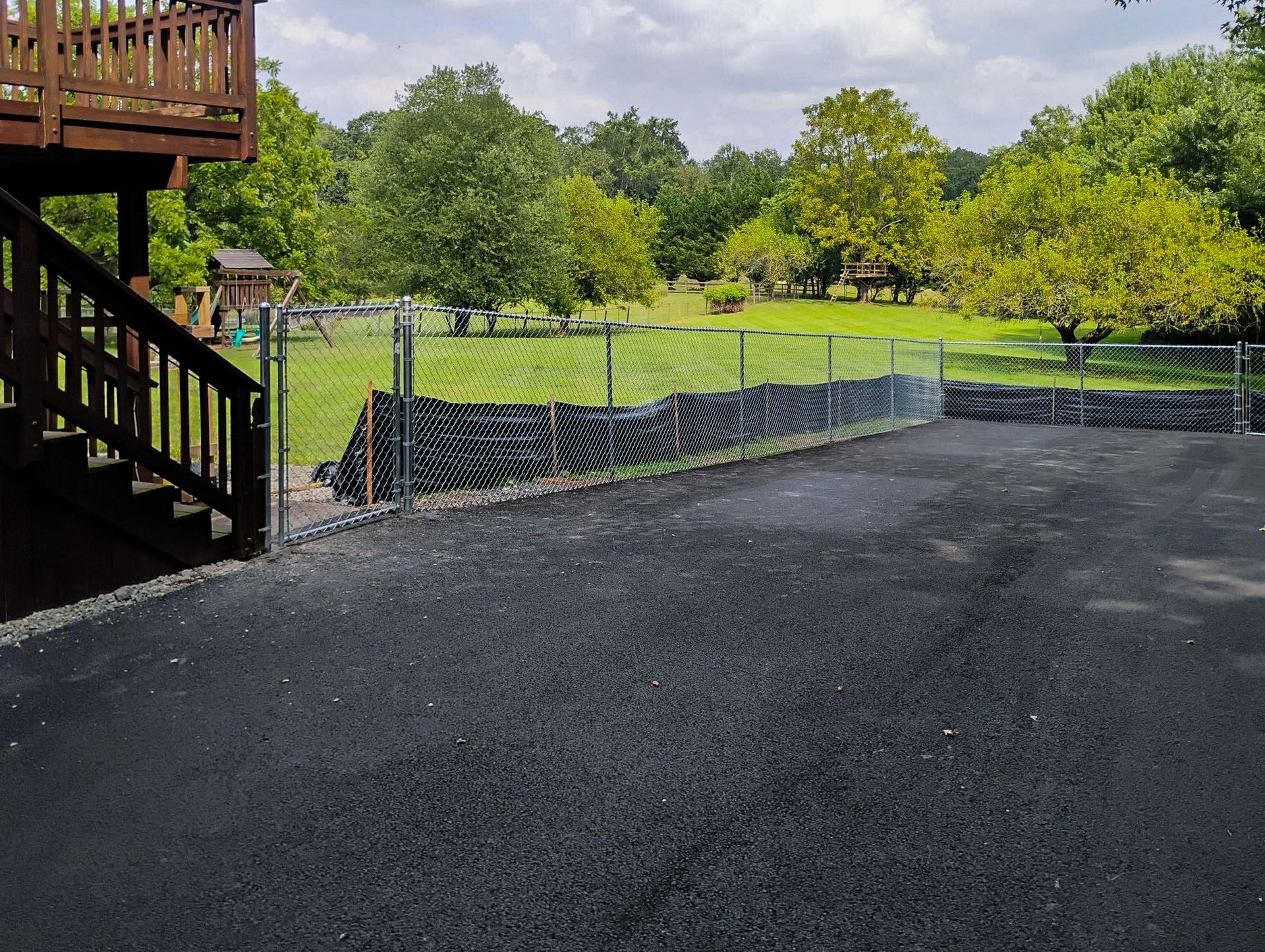 Asphalt driveway leading to chain-link fence bordering a grassy area with trees and a deck.