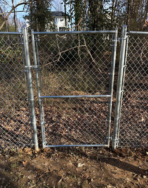 Chain-link fence with gate, set in dirt. Gate is open, leading to overgrown vegetation and a house in the distance.
