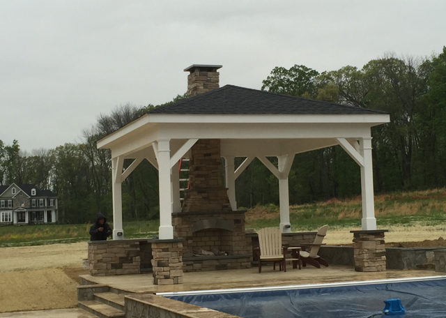 Gazebo with stone fireplace by a pool, white frame, black roof, two chairs.