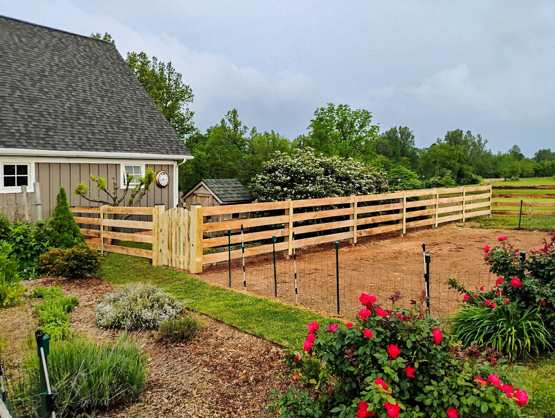 Wooden fence surrounds a garden with a house, bushes, and cloudy sky.