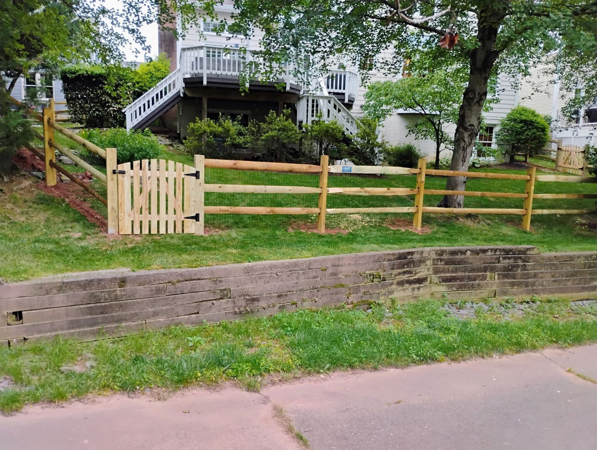 Wooden split-rail fence with a gate, in front of a house on a grassy hill, with a retaining wall in the foreground.