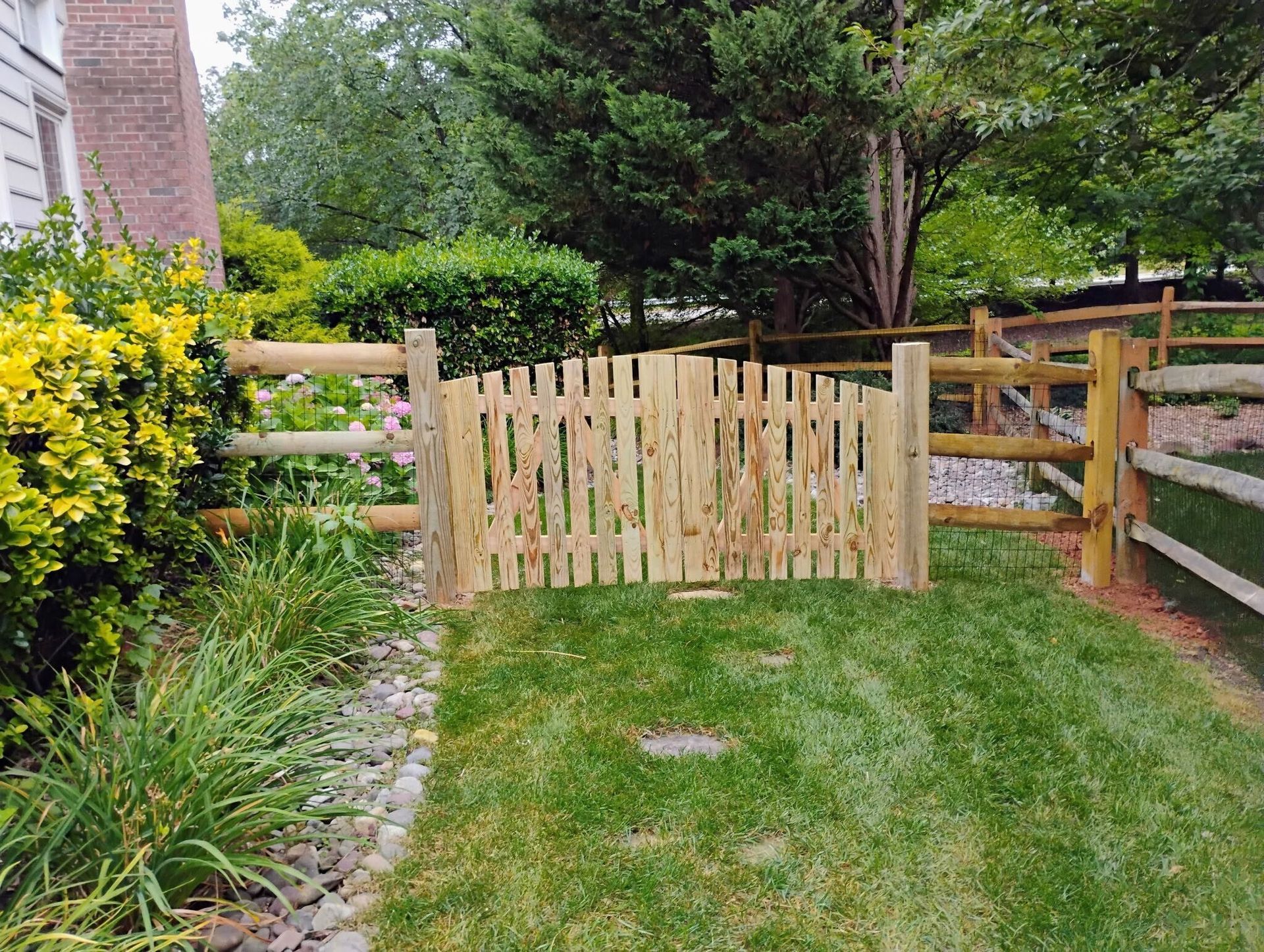Wooden gate in a grassy yard, surrounded by split-rail fence and greenery.