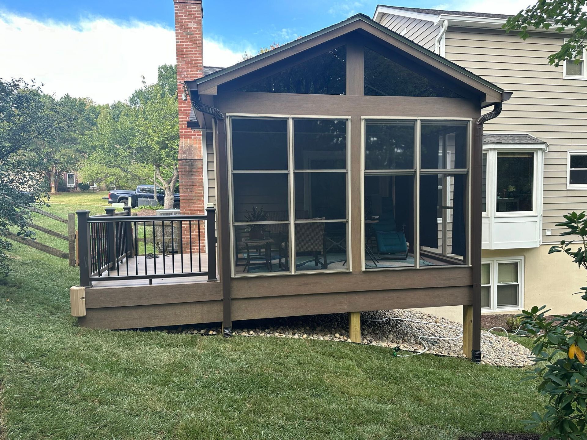 Screened porch with brown siding, connected to a house with a deck and black railing, set on a grassy hill.