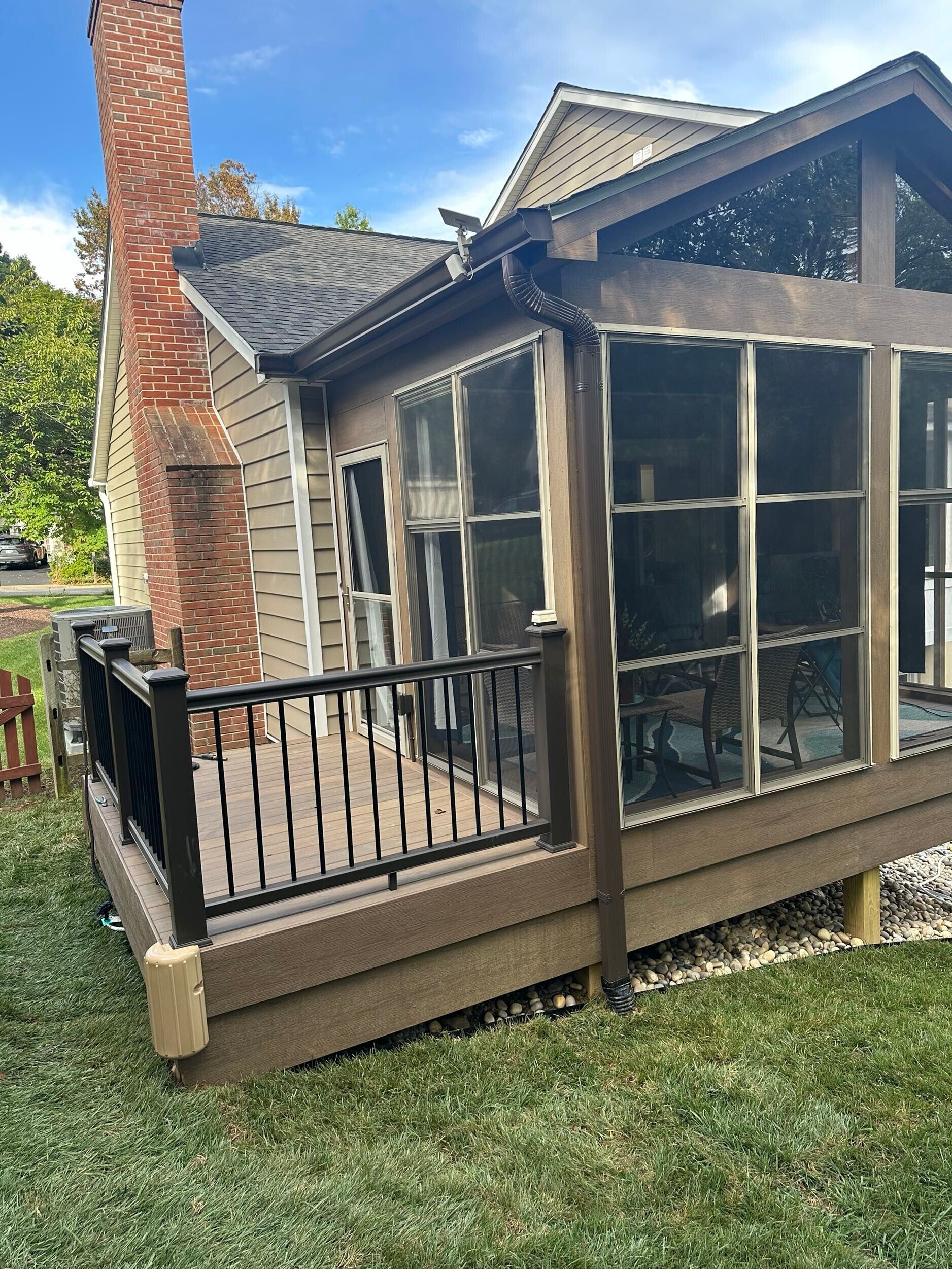 A deck with black railing and brown siding, attached to a house with a sunroom and brick chimney.