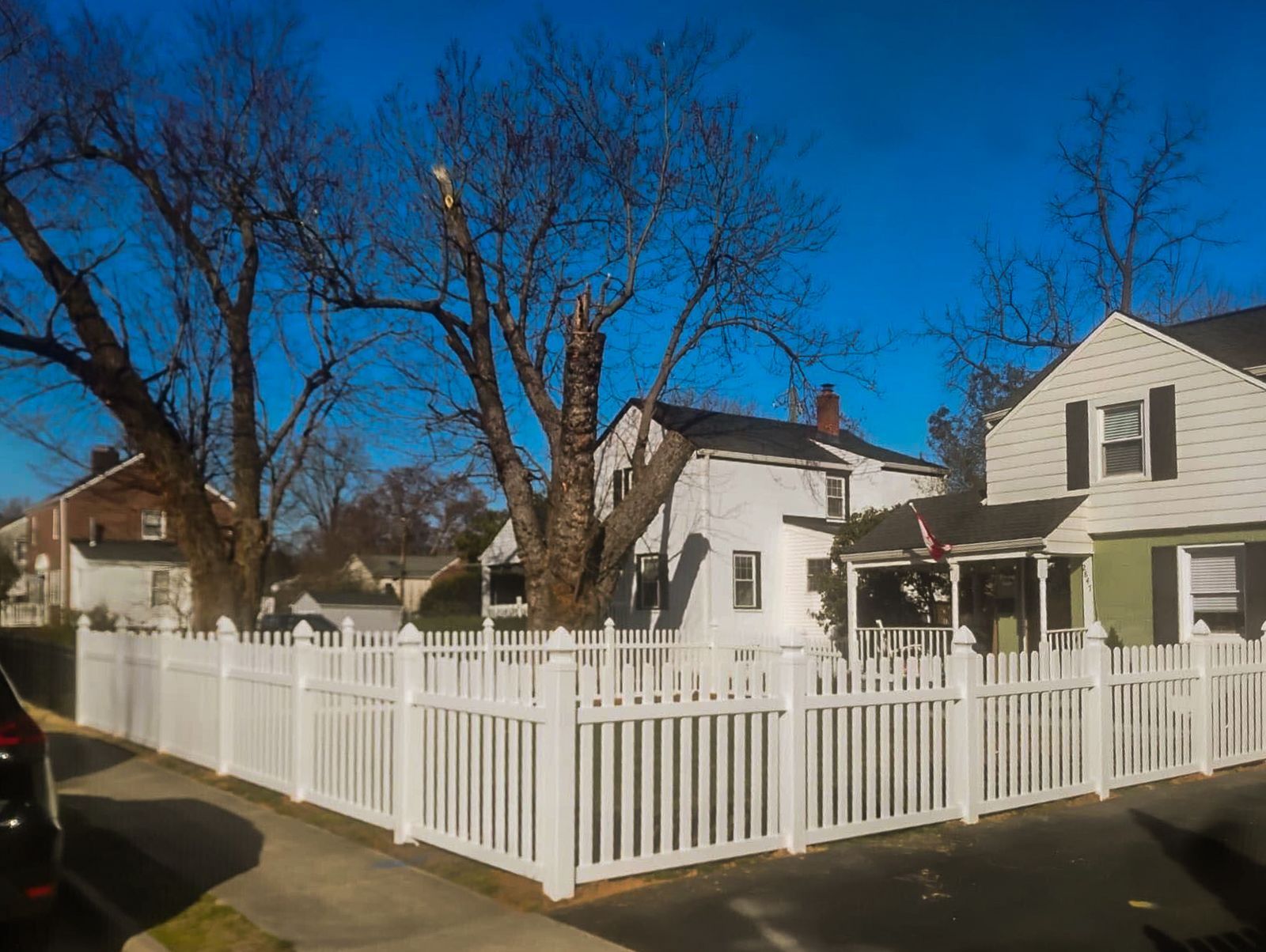 White picket fence surrounding a yard with a house and bare trees against a blue sky.
