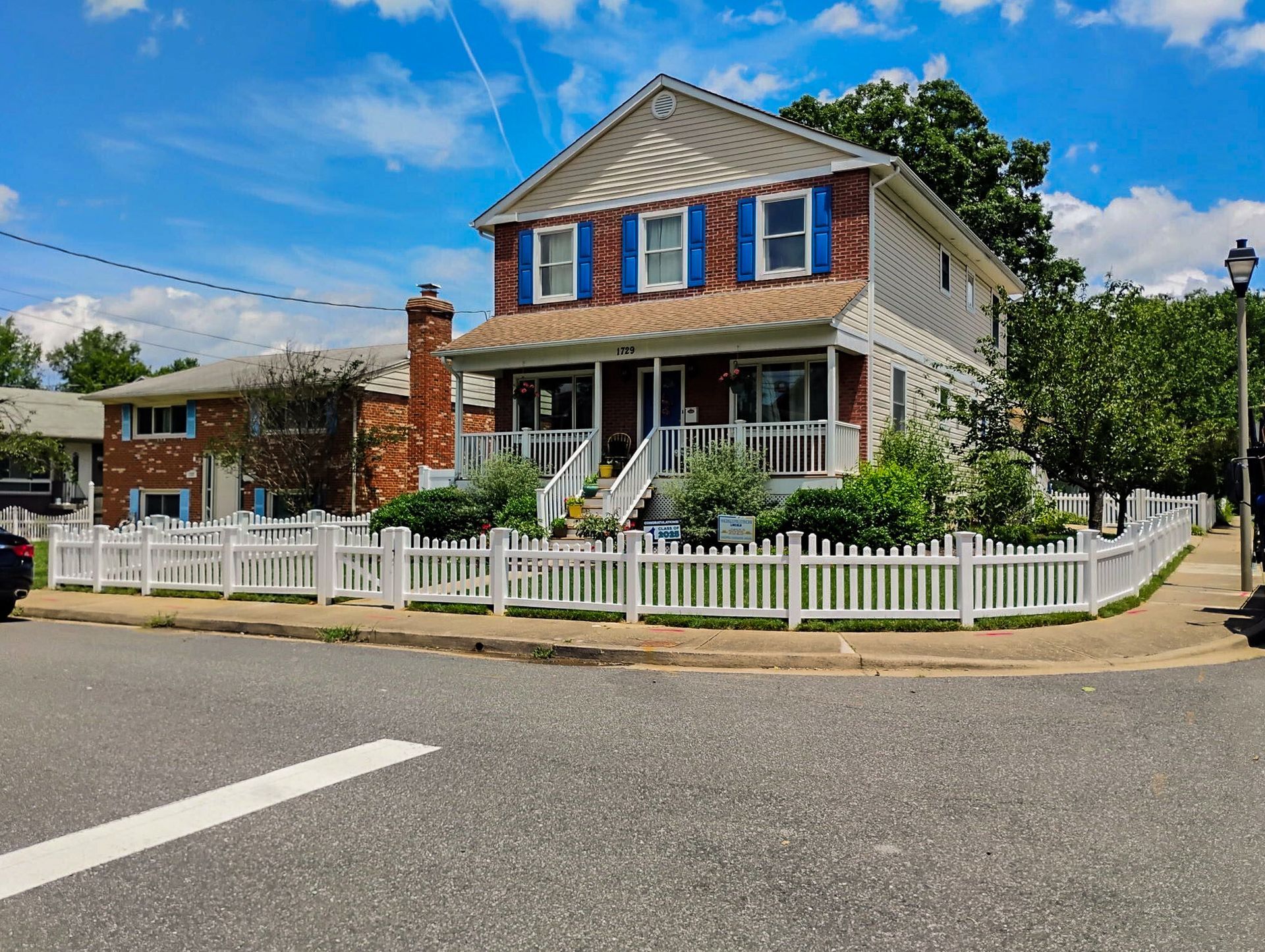 Two-story house with white picket fence on a corner lot, blue shutters, brick chimney, and clear blue sky.