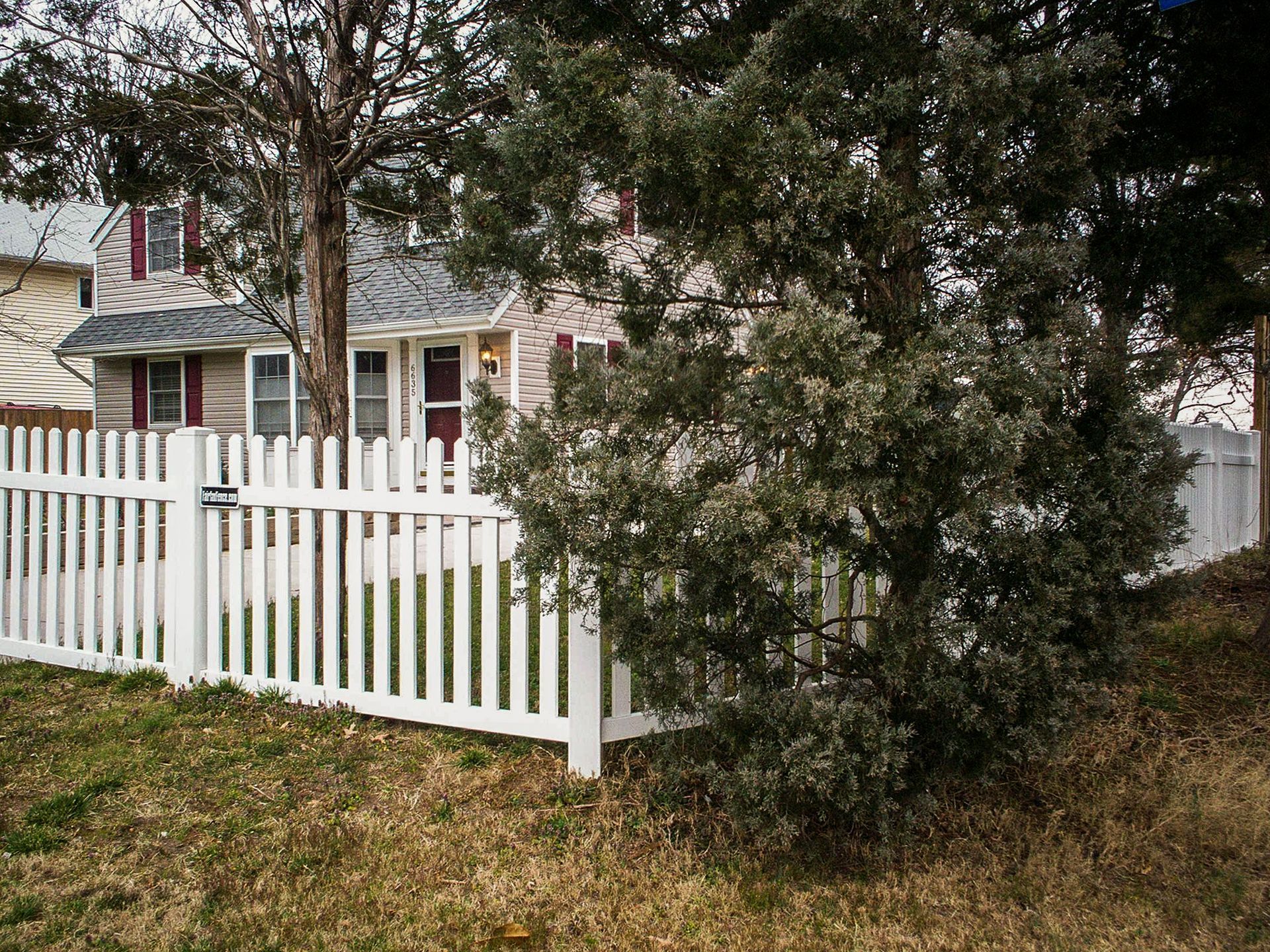 White picket fence in front of a house. Shrubbery and trees obscure some of the house.