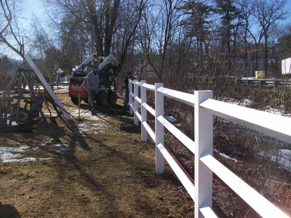 Workers installing a white post-and-rail fence with a tractor on a sunny day near trees.