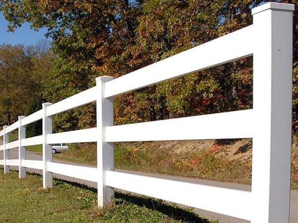 White three-rail fence alongside a road, autumn trees in the background.
