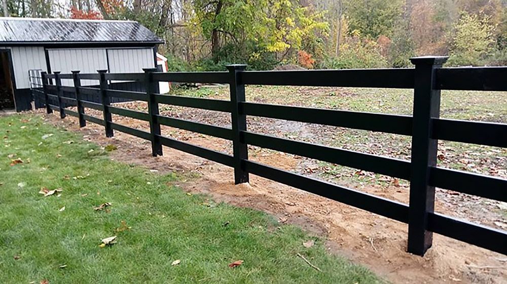 Black wooden fence bordering a grassy area and a gravel path, with a building and trees in the background.