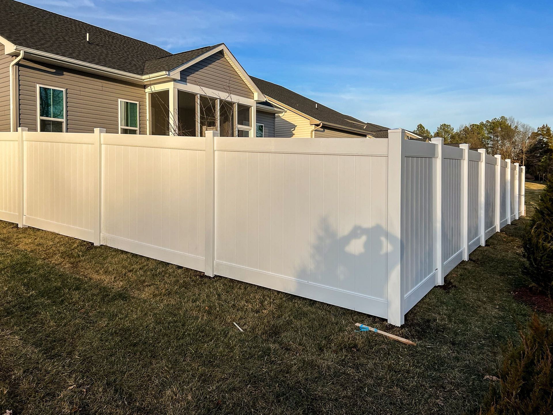 White vinyl fence surrounding a house with a gray roof, in a grassy yard, under a blue sky.