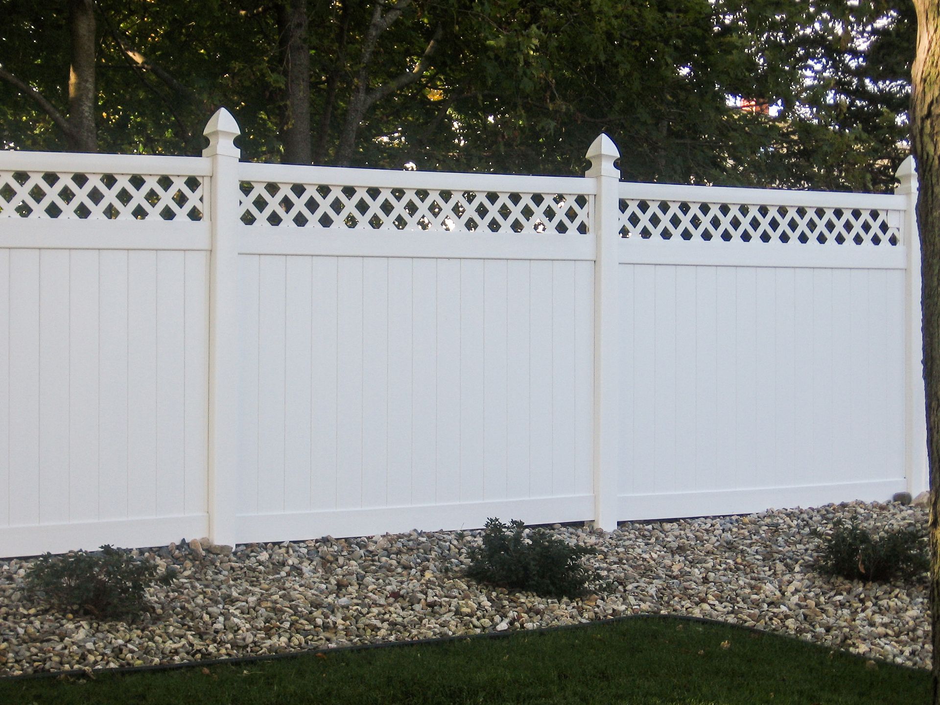 White vinyl fence with lattice top, in a yard with gravel and green grass.