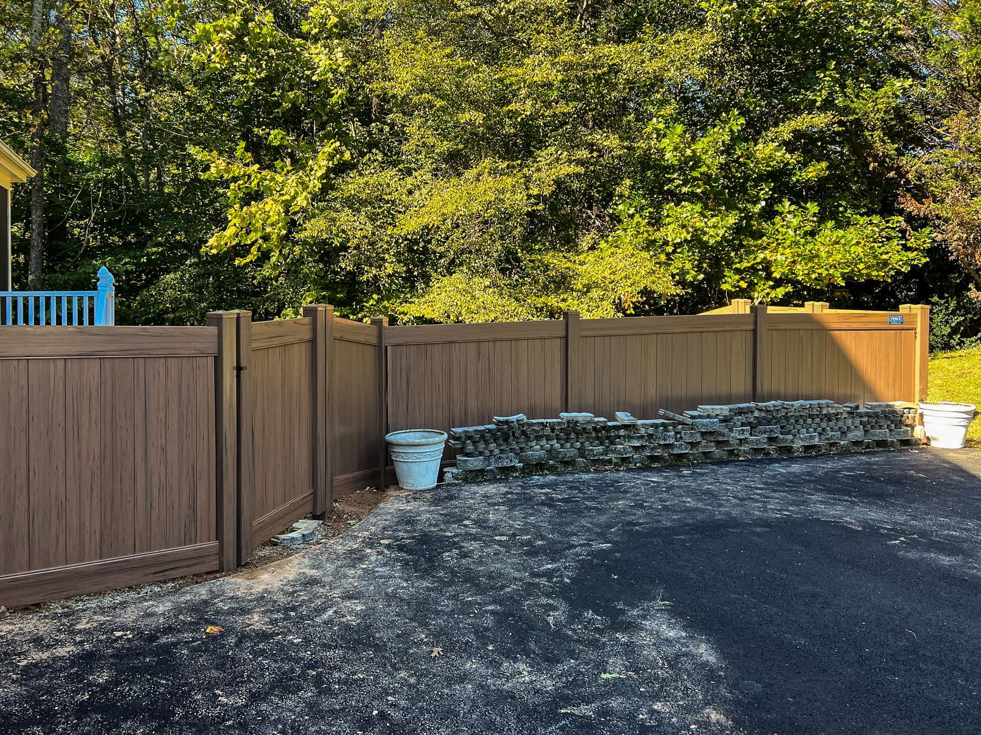 Brown fence along a driveway with trees in the background, two white buckets near a stacked stone wall.
