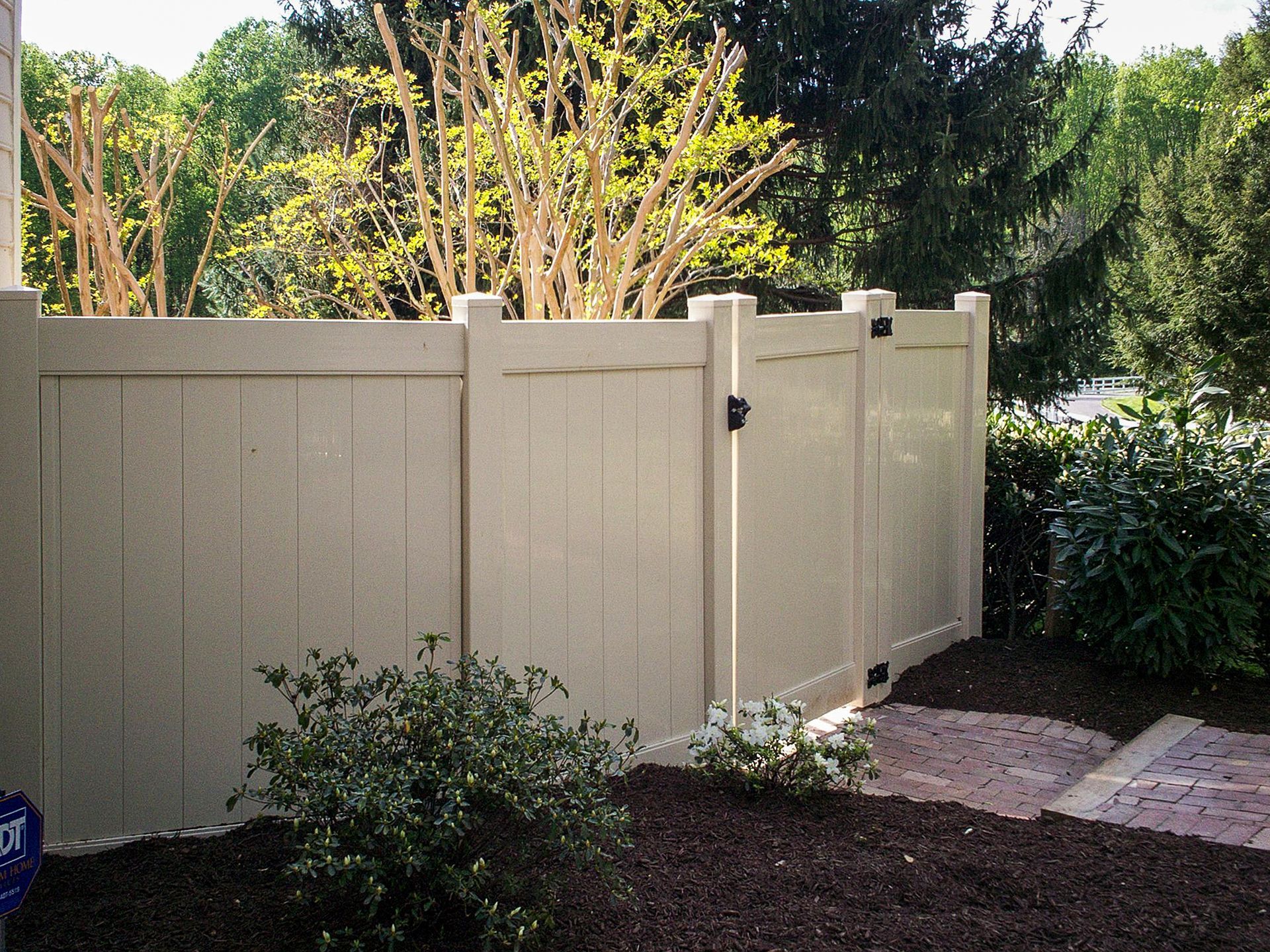 Beige vinyl fence with gate in front yard with shrubbery and trees.