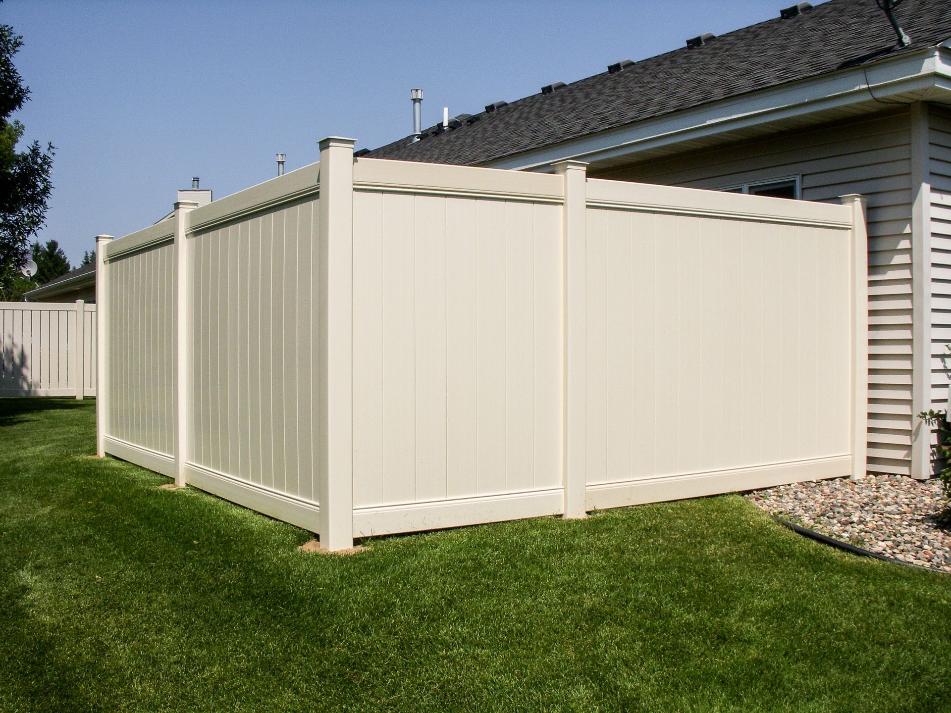 Beige vinyl fence enclosing a green lawn next to a house with a dark roof.