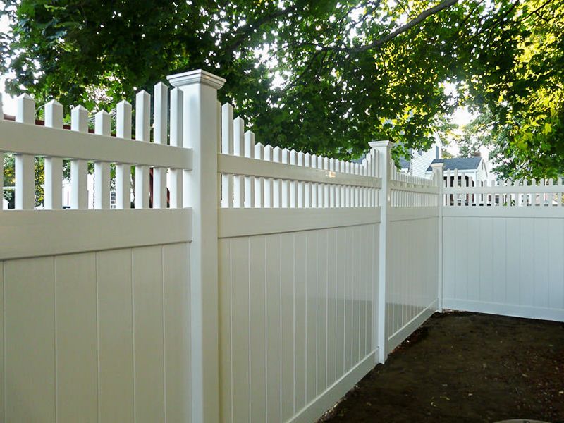 White picket fence in a residential setting, with a vertical panel design and a green, leafy backdrop.
