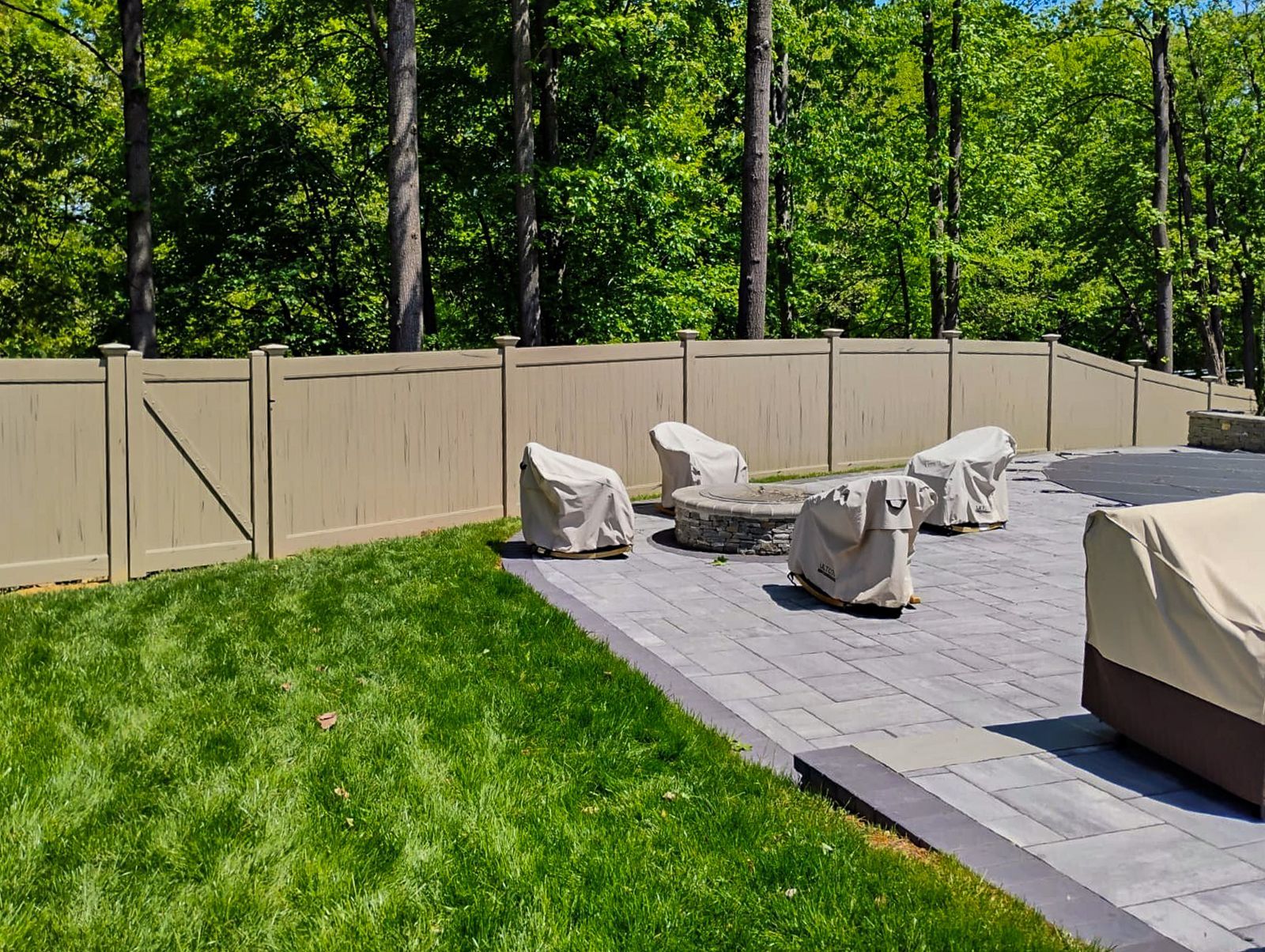 Backyard patio with covered furniture, a fire pit, and a tan fence against a backdrop of trees.