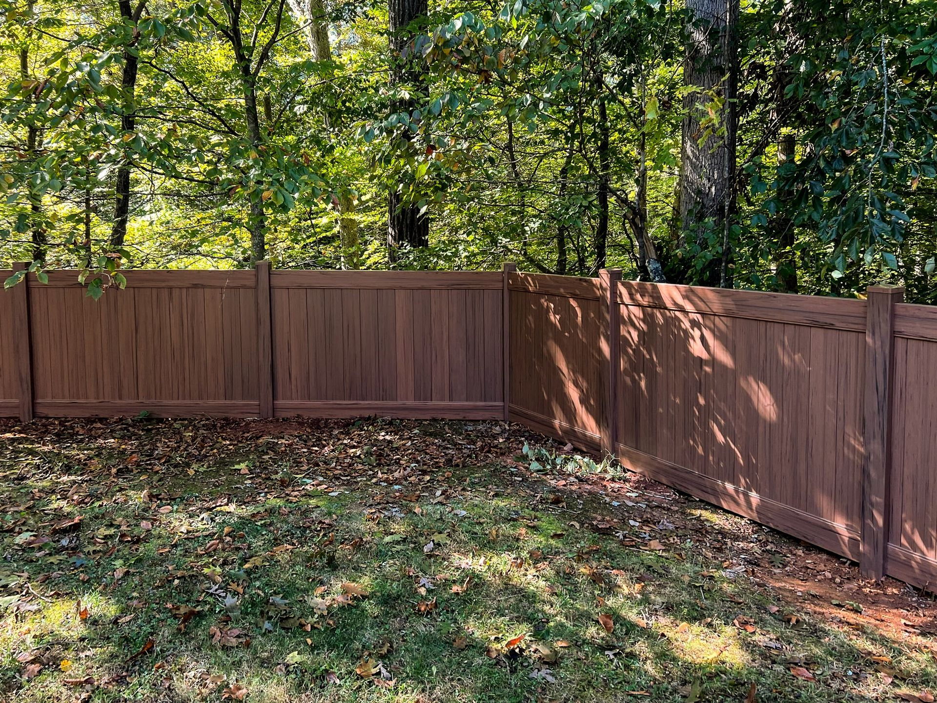 Brown wooden fence encloses a backyard with trees and fallen leaves in the grass.