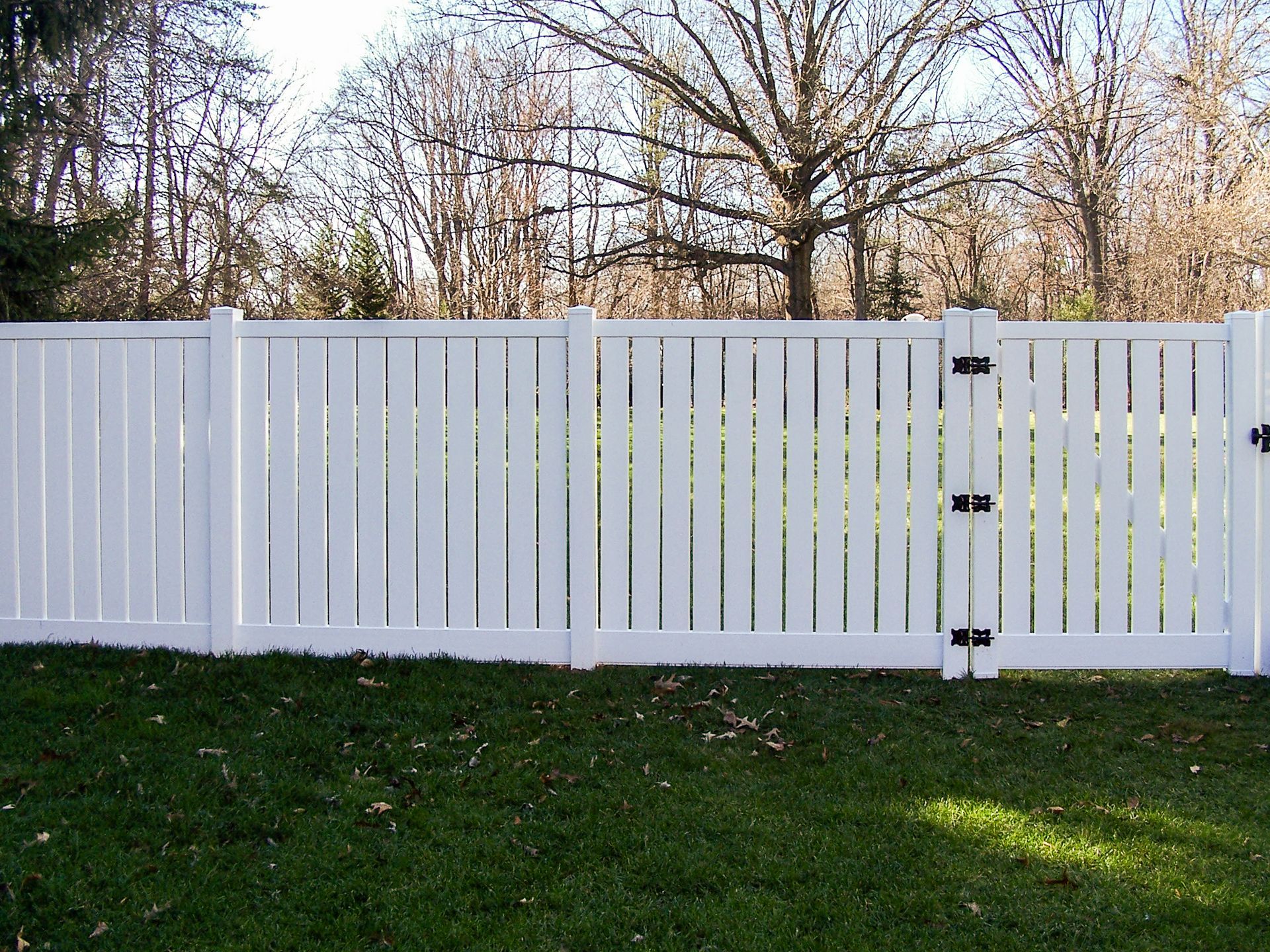 White picket fence with a gate in front of a grassy yard with trees in the background.