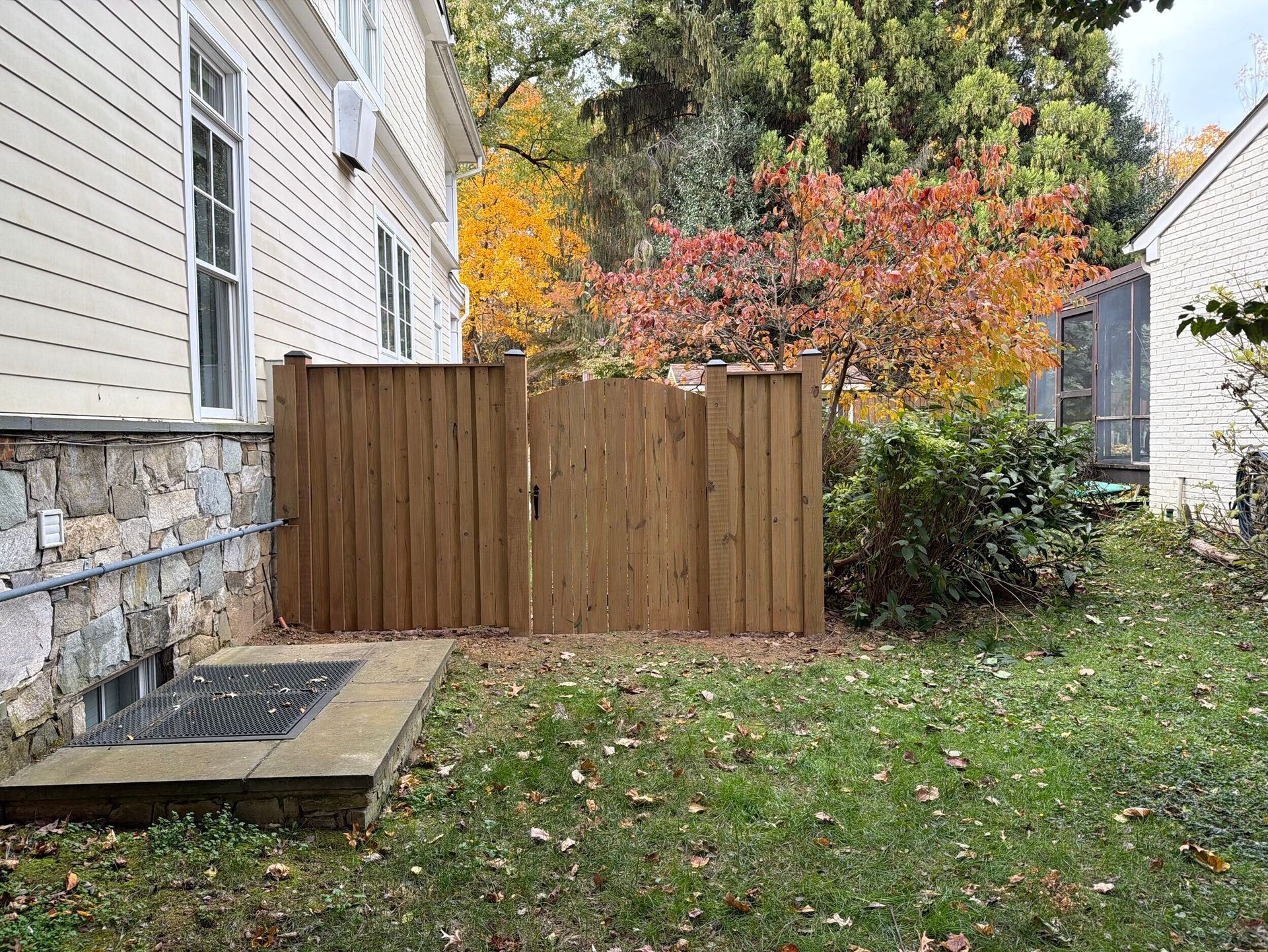 Wooden fence beside a house with autumn foliage in the background. Grass and a window well in the foreground.