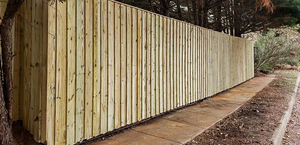 Wooden fence along a concrete path, next to trees and brush.