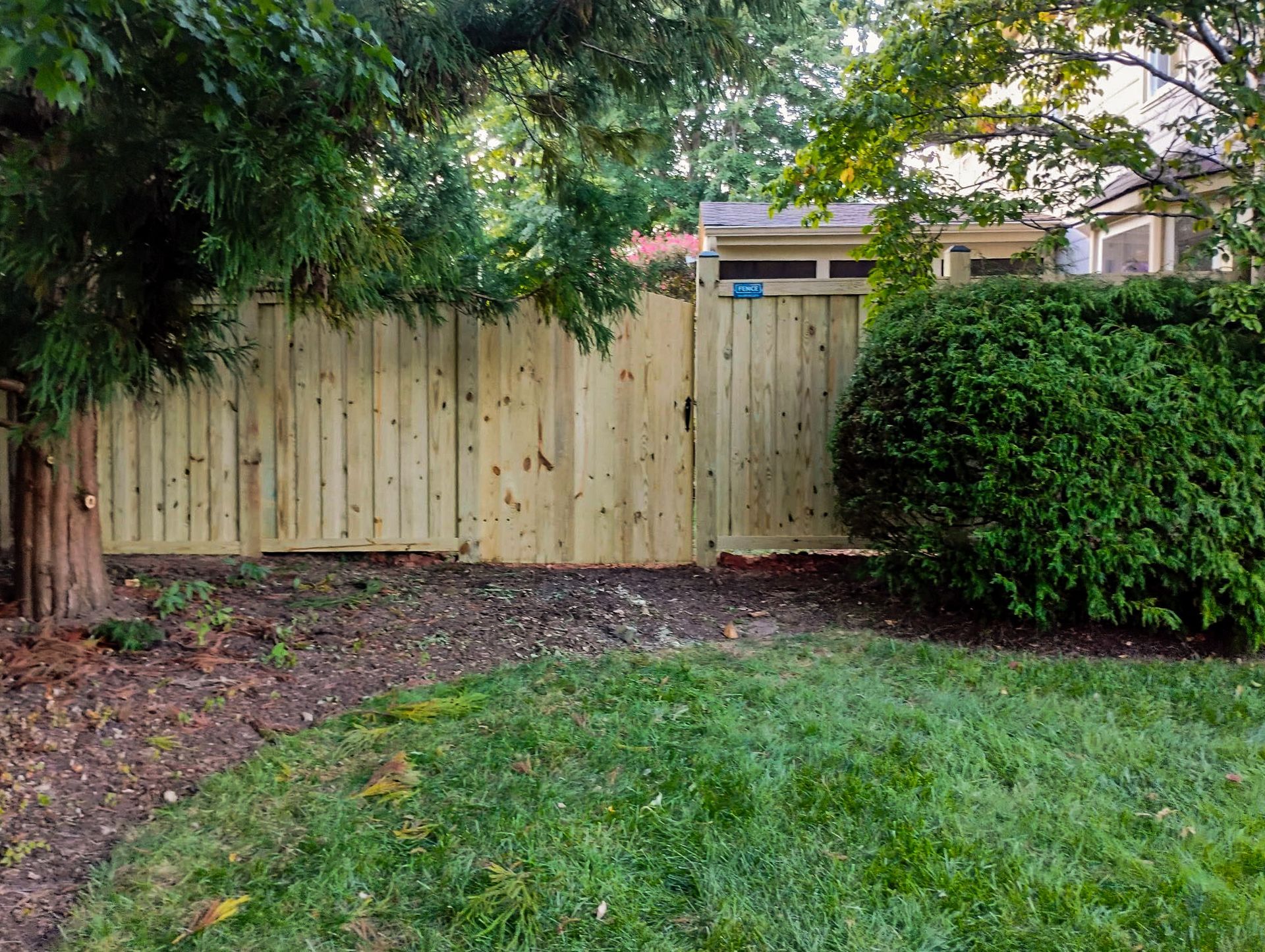 Wooden fence with gate, bordered by green grass, mulch, and trees.