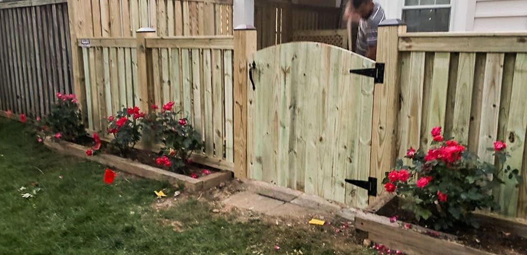 Wooden gate in a wooden fence with red flowers in a garden bed. A person is visible in the background.