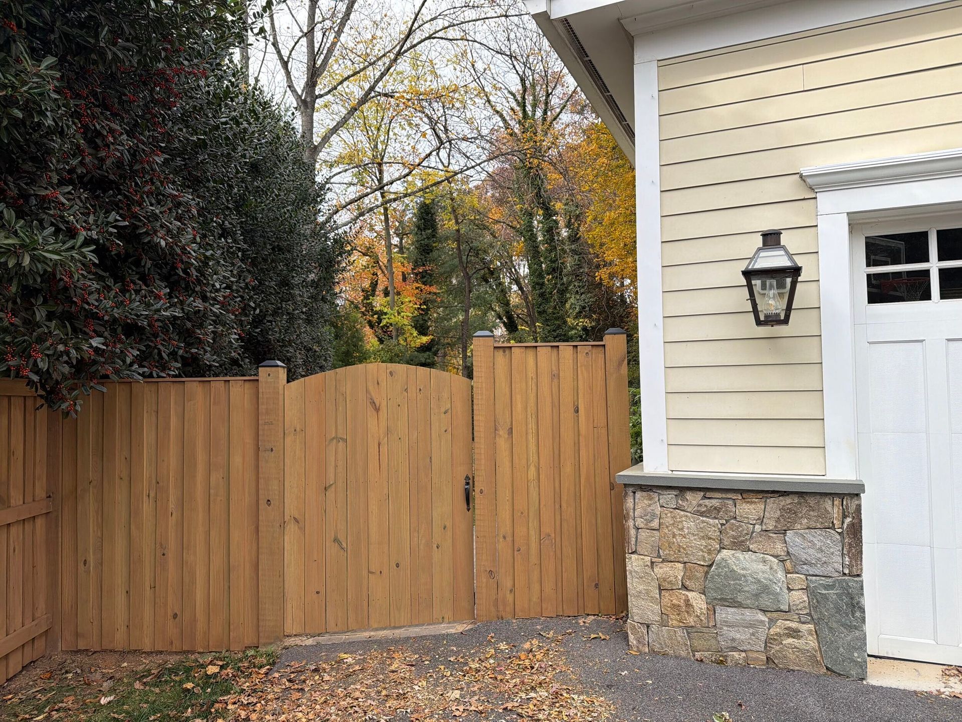 Wooden fence with gate next to a garage with tan siding, stone base, and a lantern.