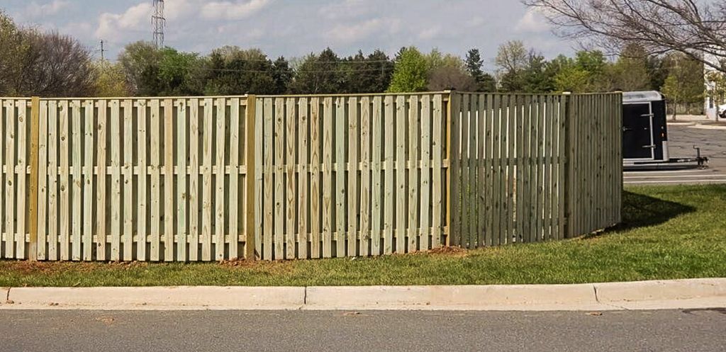 A wooden fence borders a grassy area next to a road, with trees and a trailer visible in the background.