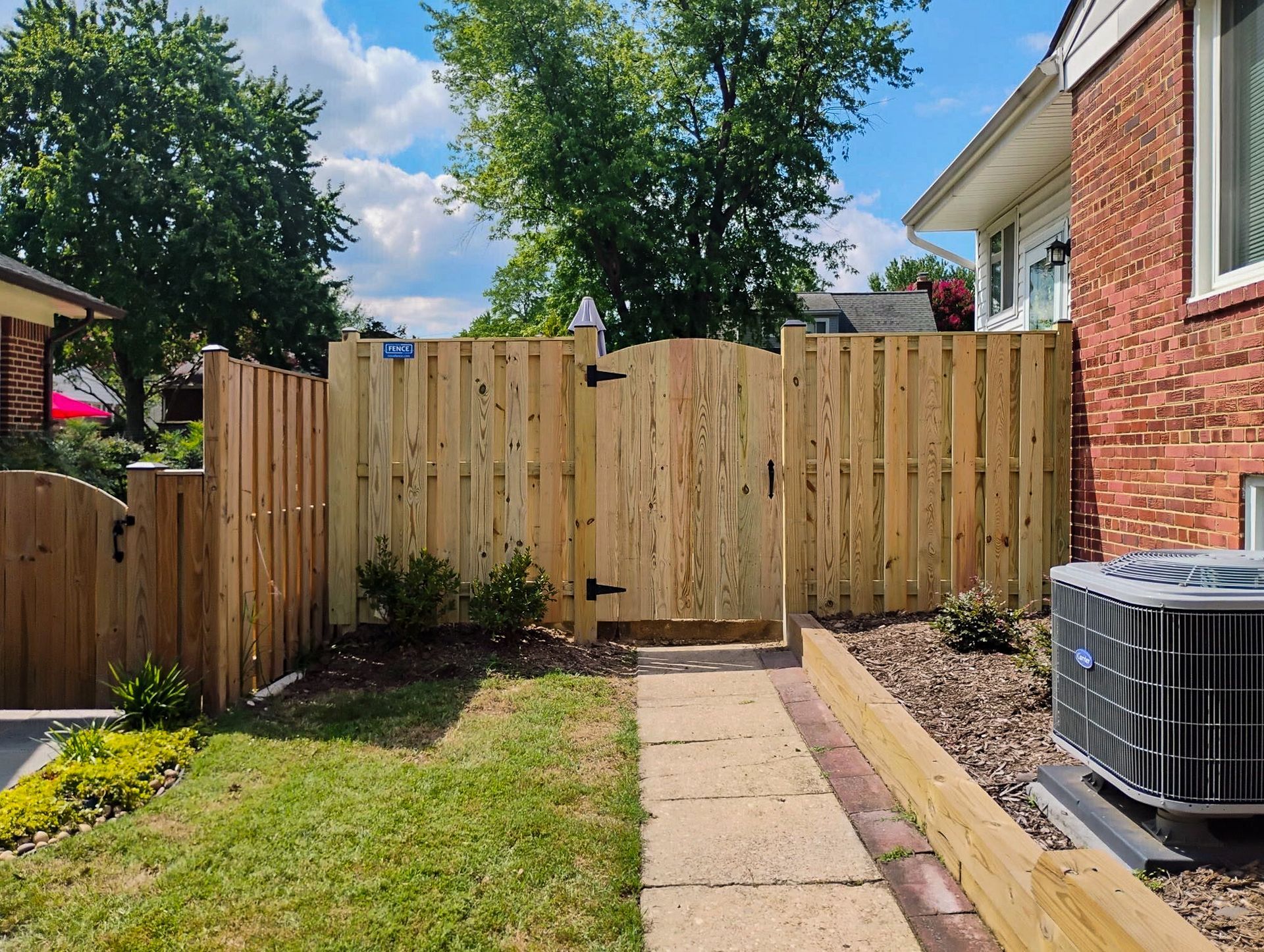 Wooden fence with gate in front of a brick house. Pathway leads to the gate. Green grass and blue sky.