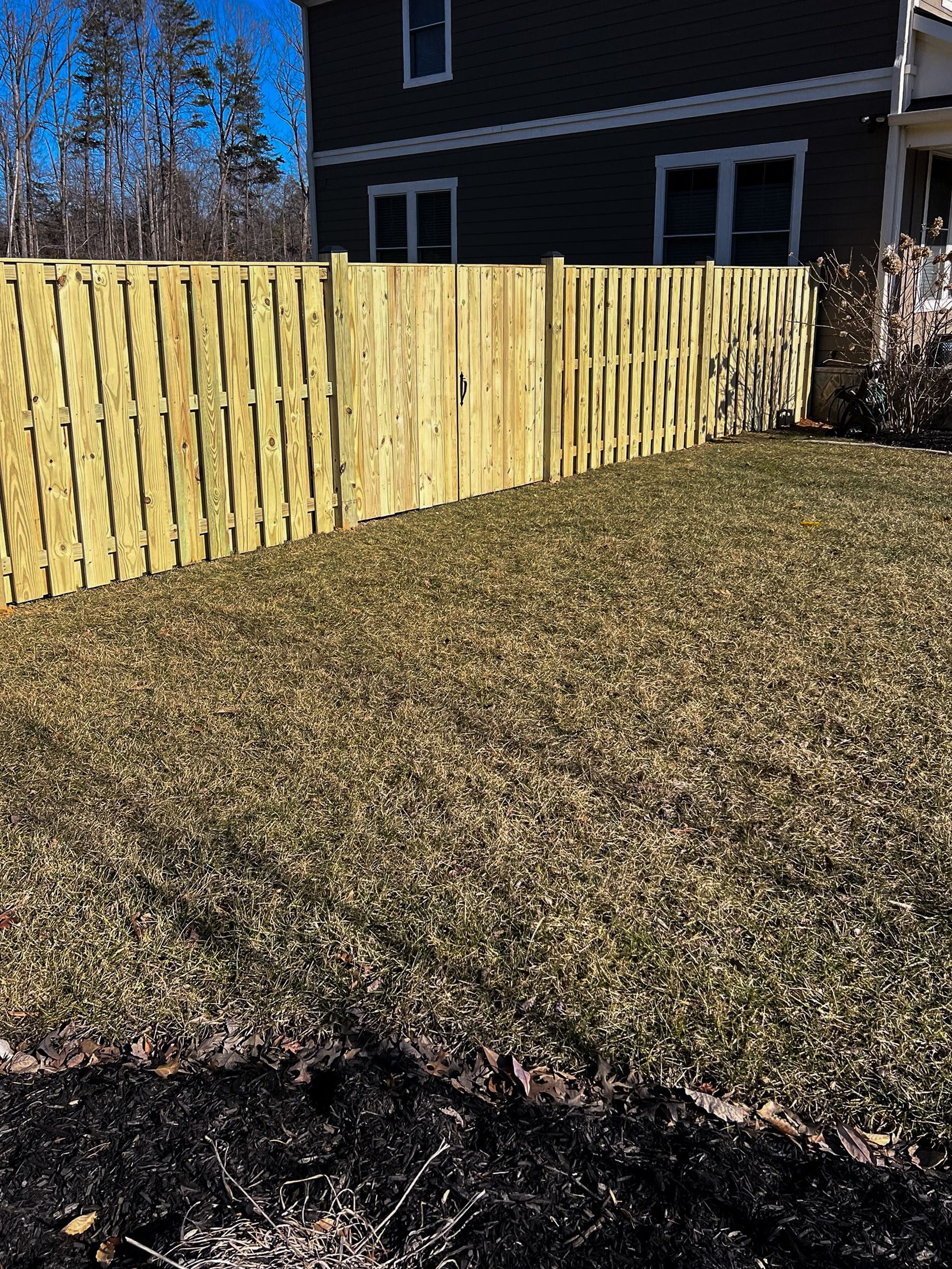 Wooden fence bordering a grassy yard with a house in the background on a sunny day.