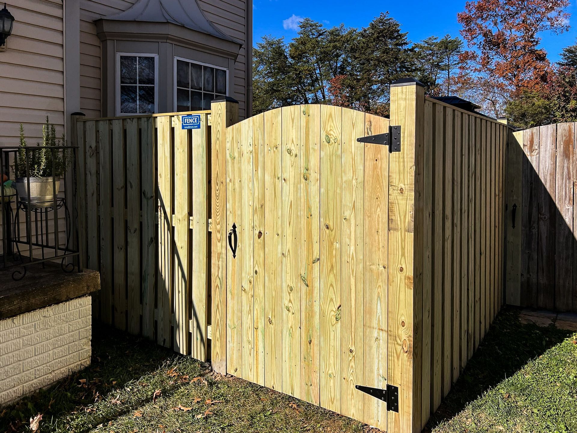 Wooden gate in a backyard, flanked by fences. Gate has black hardware and a curved top.