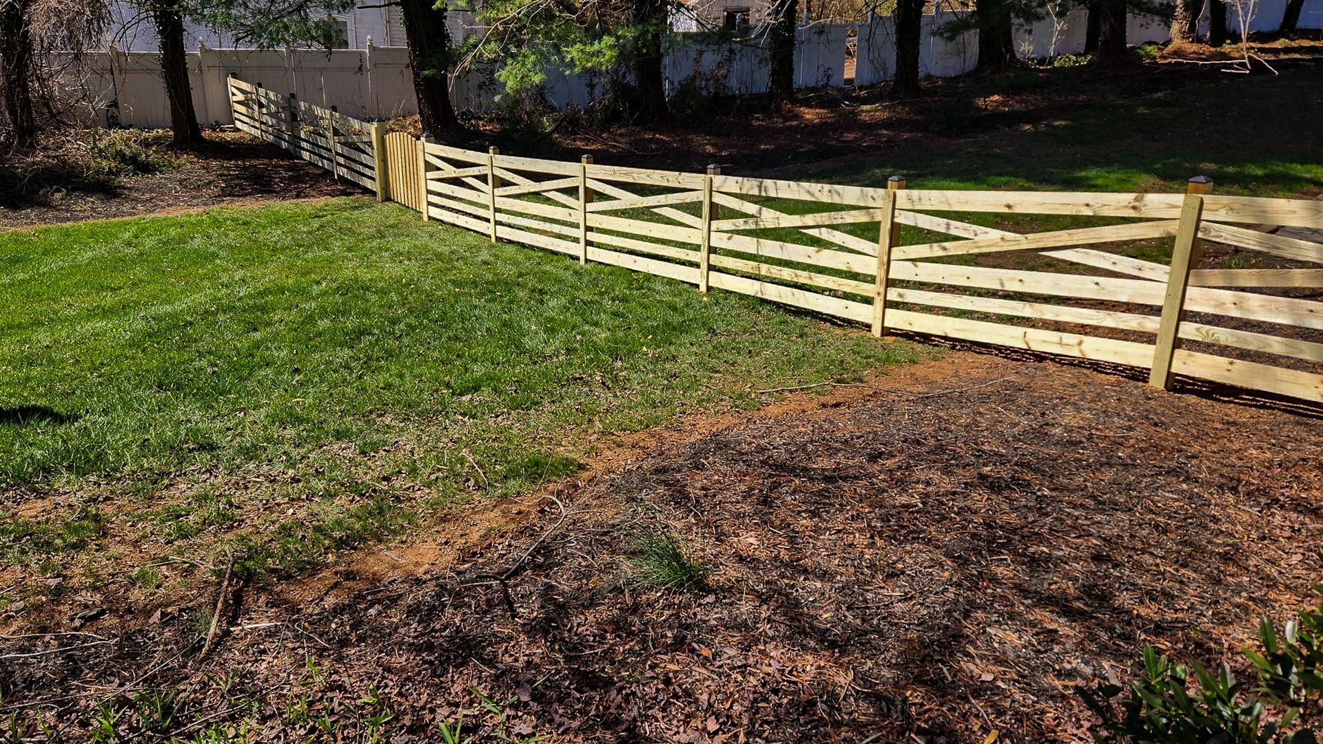 Wooden fence bordering a yard with green grass and brown mulch. Trees and a white fence are in the background.