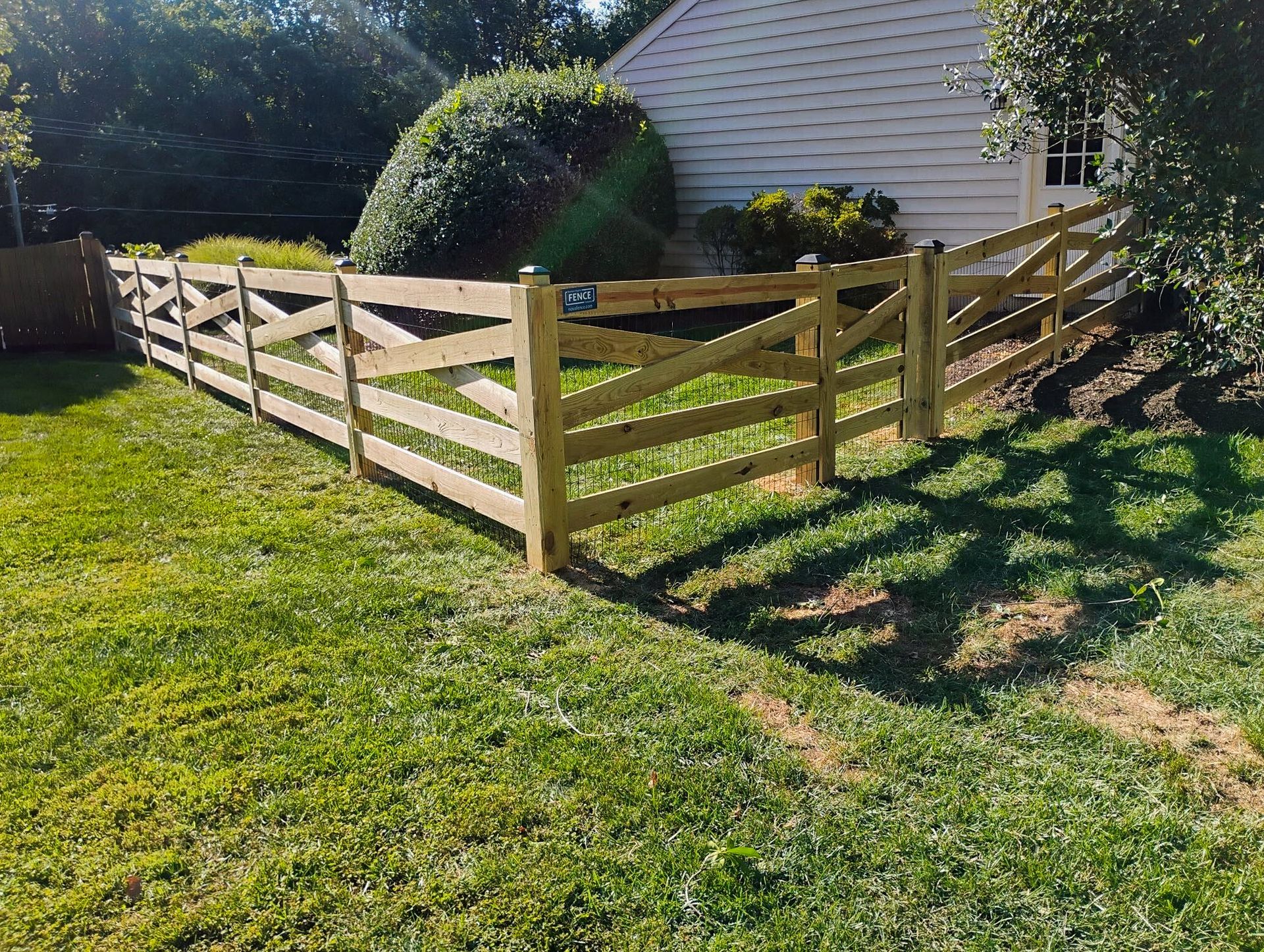 Wooden fence around a grassy yard, beside a house with a bush in the background.