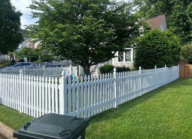 White picket fence surrounds a house with a green lawn. A green trash bin is in the foreground.