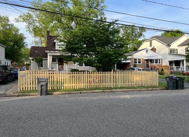 A light-colored wooden picket fence in front of a house, with street and neighboring houses visible.