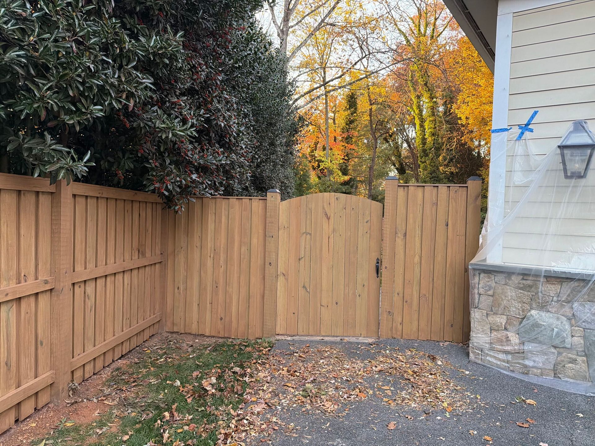 Wooden fence with gate leading to a yard with fallen leaves; autumn trees visible in the background.