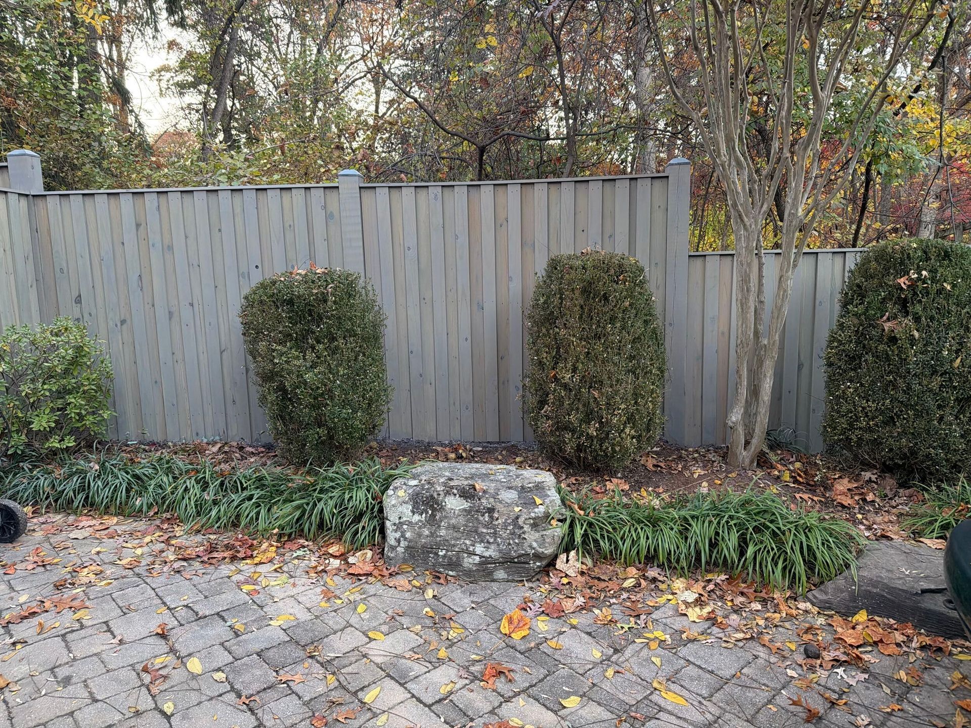 Gray fence with trimmed green bushes and a large stone on a brick patio.