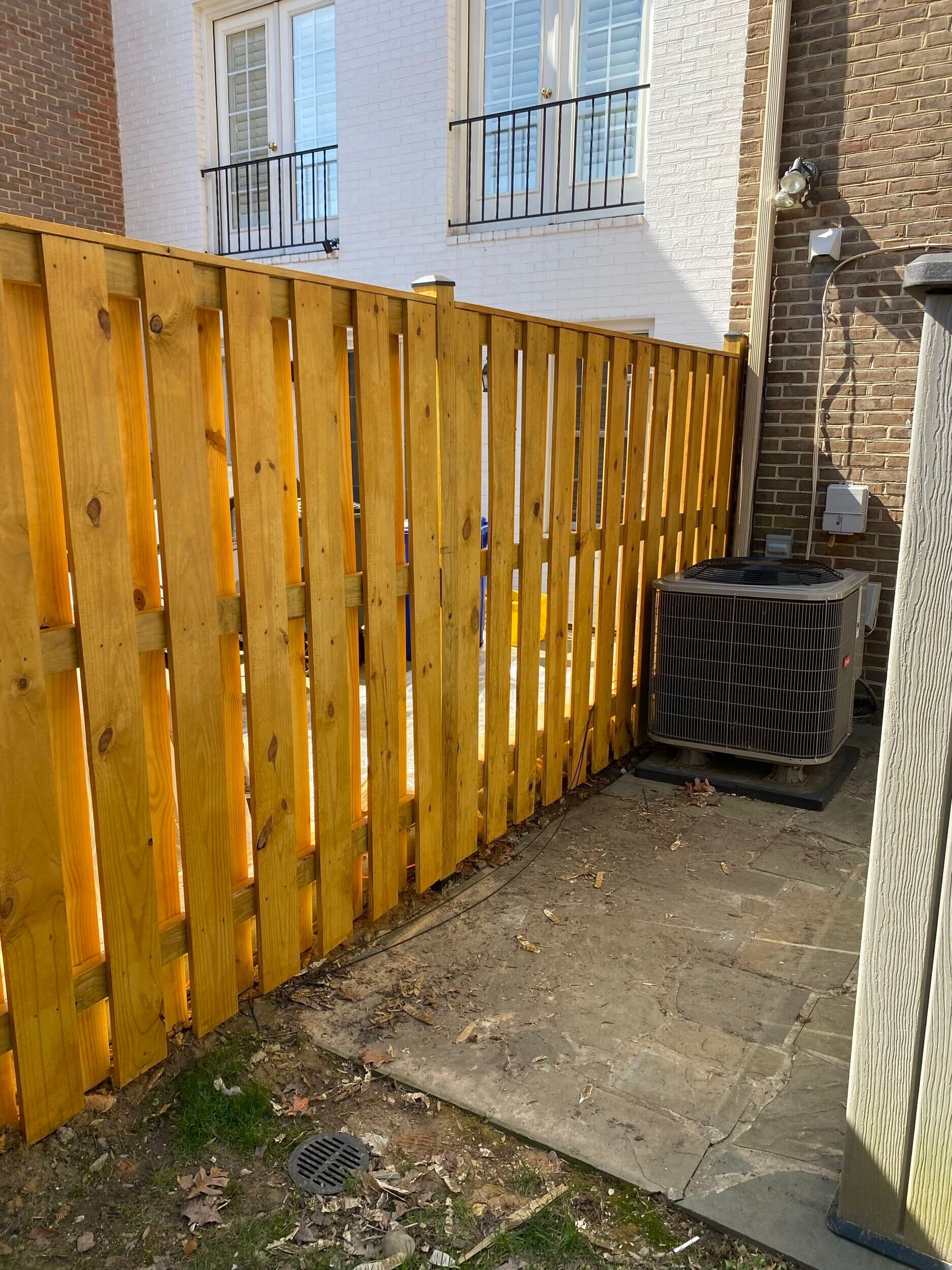 Yellow wooden fence beside a building with an AC unit on a concrete patio.