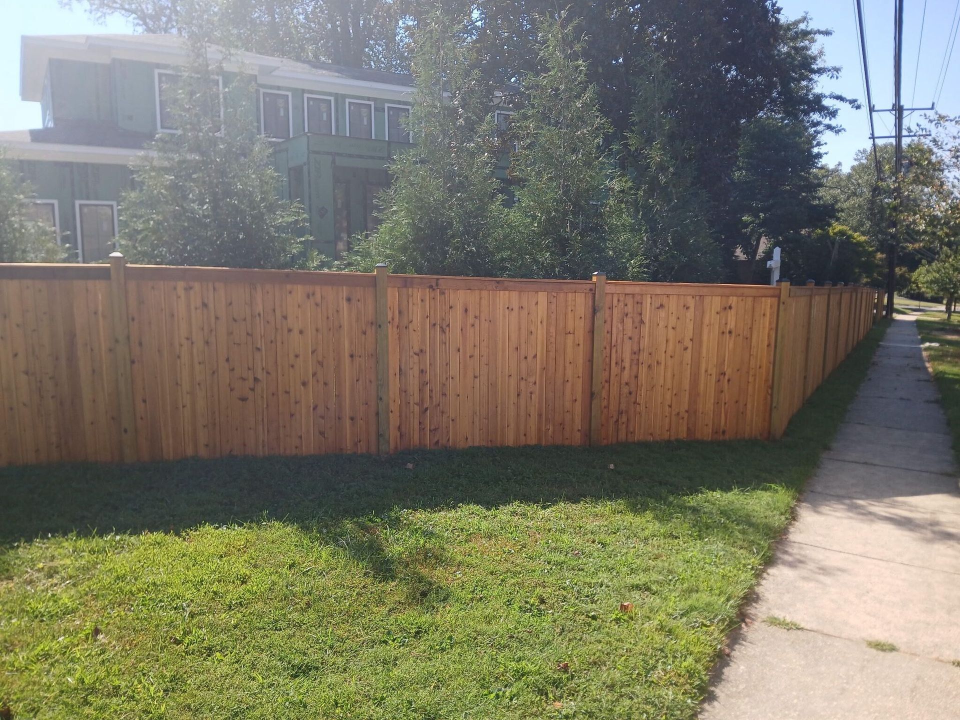 A wooden fence lines a sidewalk next to a grassy lawn in front of a house on a sunny day.