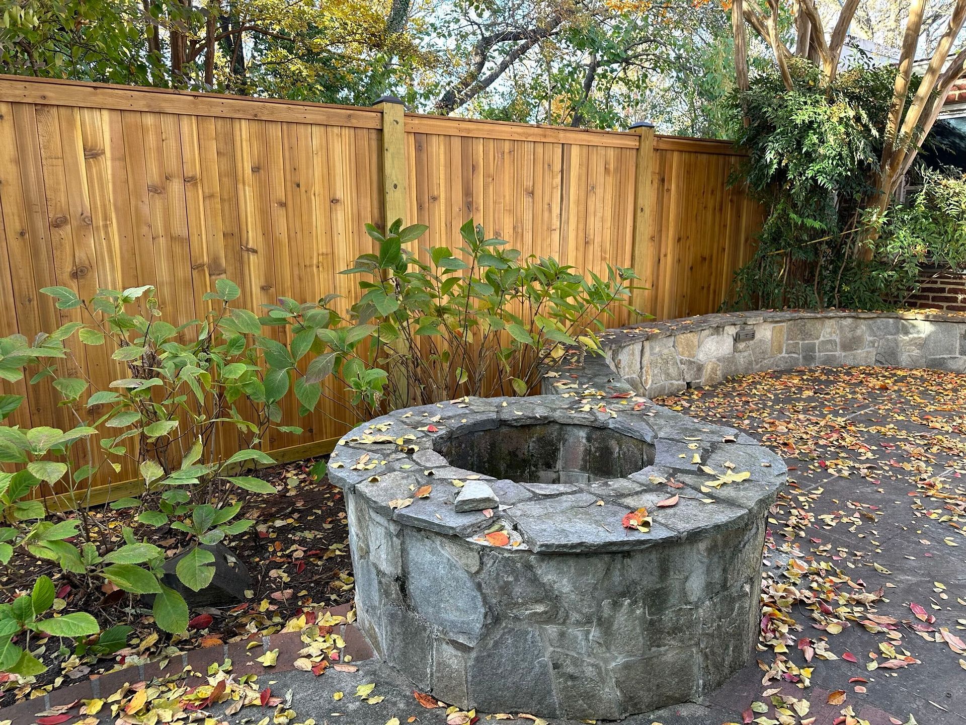 Stone fire pit surrounded by fall leaves, plants, and a wooden fence.