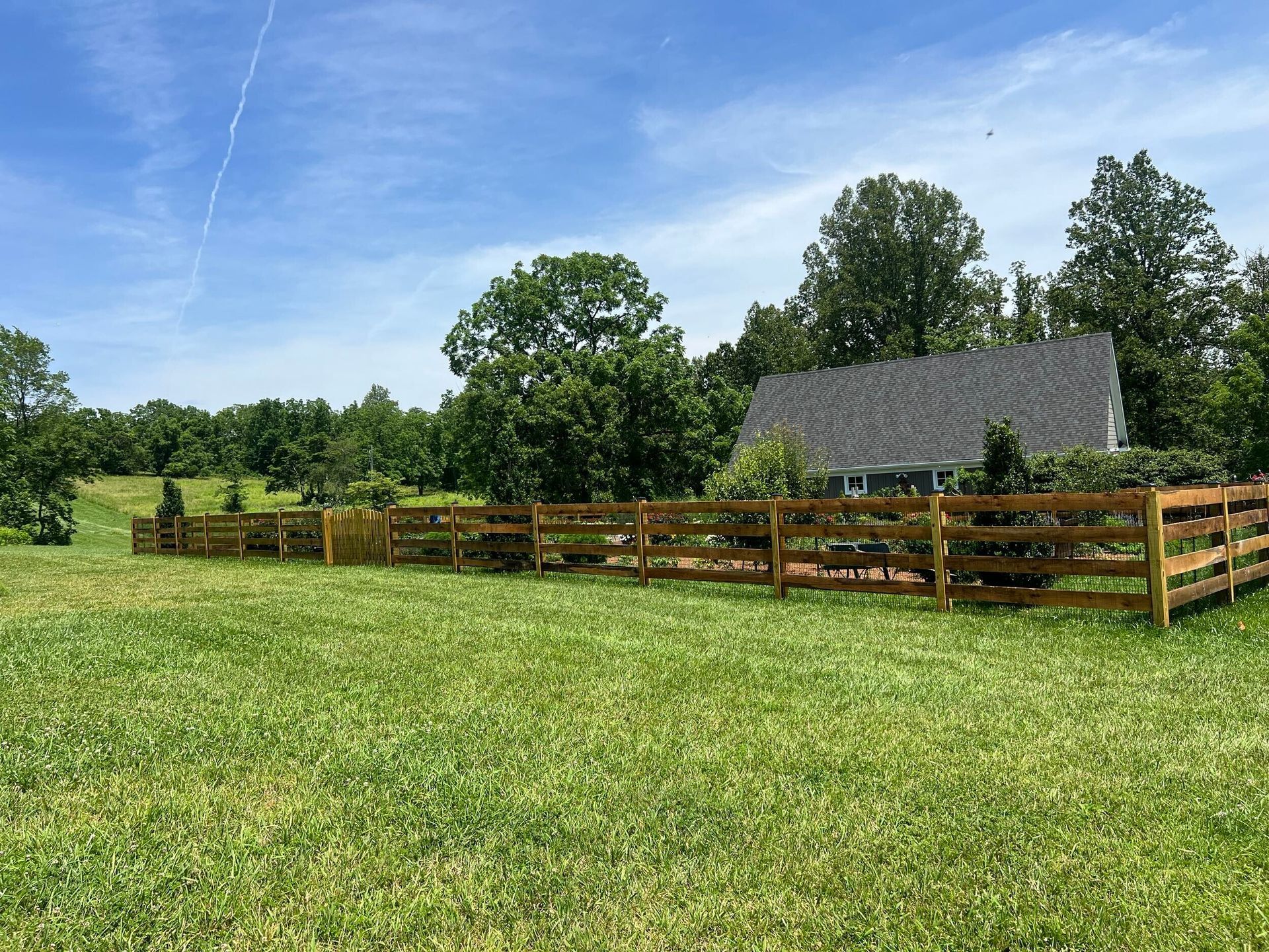 A wooden fence surrounds a grassy field, with a house and trees in the background on a sunny day.