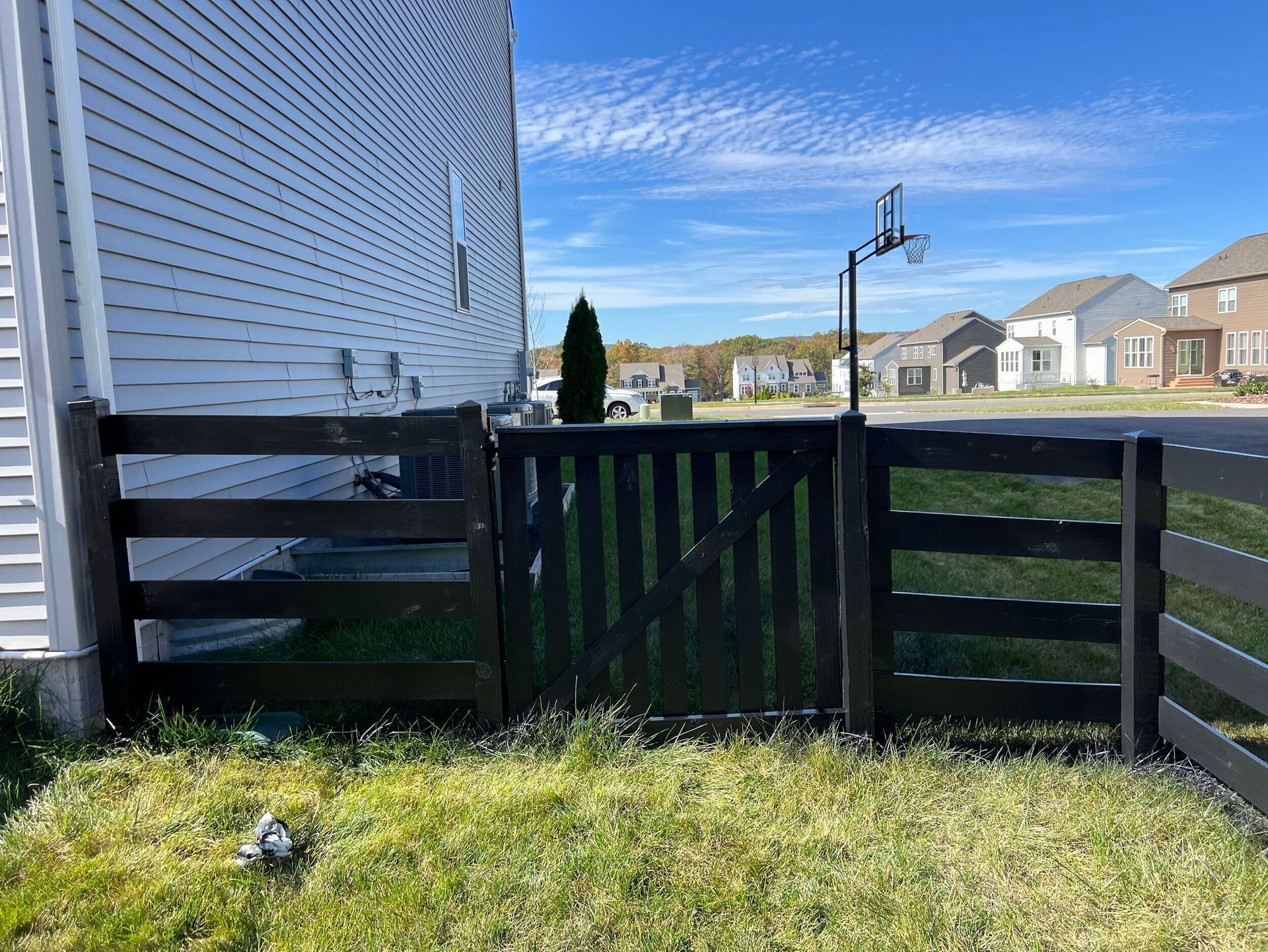 Black wooden fence with gate in front yard of a house, basketball hoop visible in the distance.