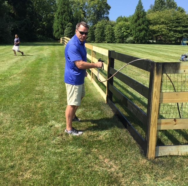 Man spray-painting a dark fence in a grassy yard, another person walking in background.