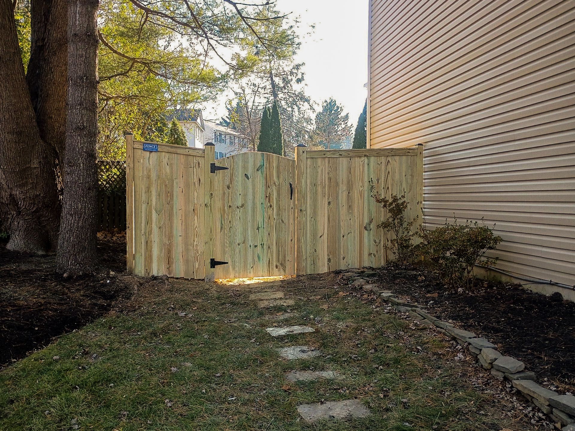 Wooden fence with a gate, next to a house and trees. A stone path leads to the gate.